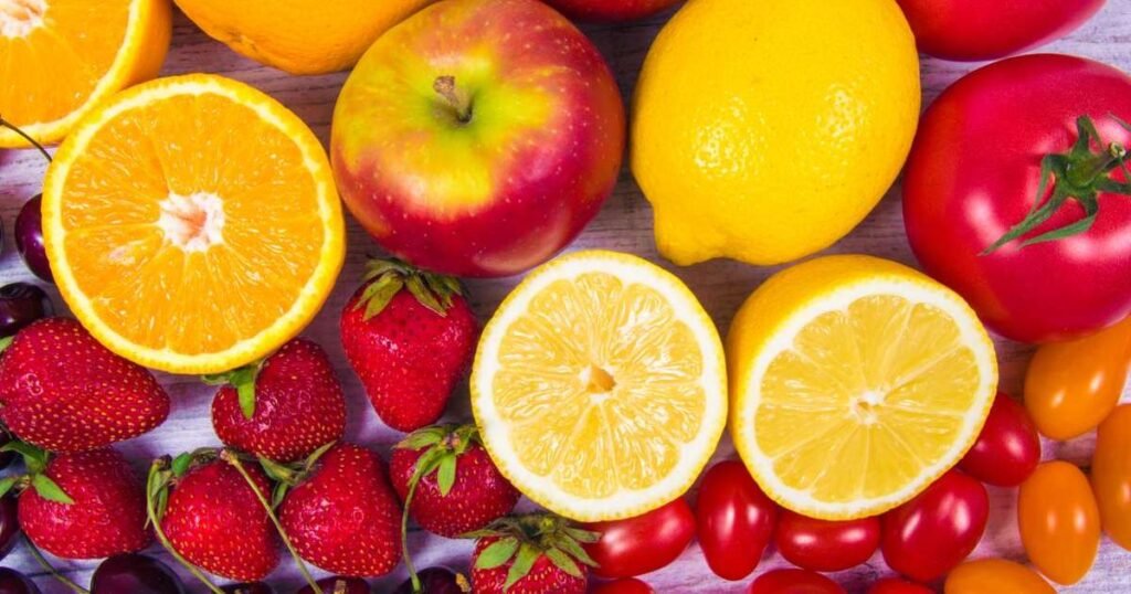 Uma variedade de alimentos ricos em vitamina C, incluindo acerola, laranja, limão, goiaba, pimentão verde, brócolis, couve, tomate, morango e kiwi dispostos em uma mesa.
